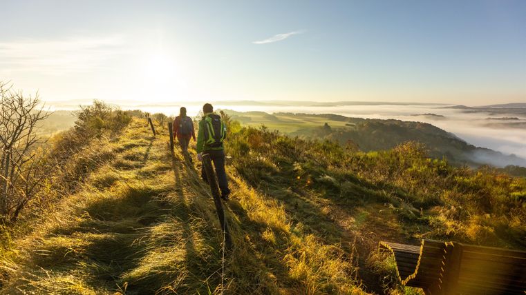 Deux randonneurs marchent le long d'un sentier à travers un paysage vallonné. En arrière-plan, un ciel dégagé et un banc de brume sont visibles.