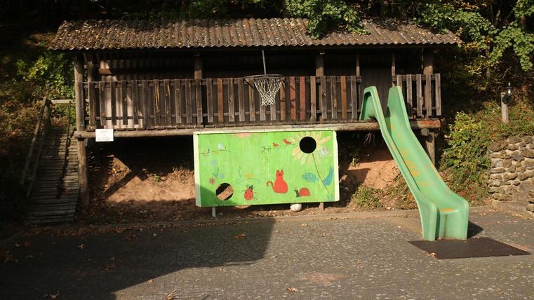 Ein Spielplatz mit einem Holzhaus und einer Rutsche. An der Wand sind farbenfrohe Bilder von Tieren und eine Basketballwurfstelle zu sehen.