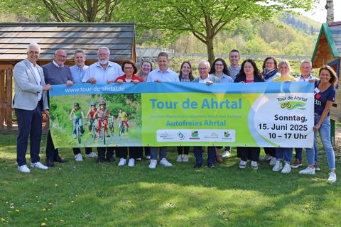 16 Personen stehen dicht beieinander, halten ein Banner für den Radaktionstag Tour de Ahrtal am Sonntag, 15. Juni 2025 hoch und lächerln in die Kamera.