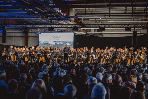 Ein prall gefüllter Raum aus Zuschauern vor einer großen Gruppe Musikanten und dem Dirigenten.