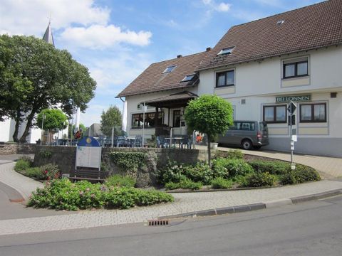 Blick von vorne auf das Gasthaus Bei Lonnen.Das zweistöckige Gebäude ist hell angestrichen und hat ein schwarzgedecktes Dach. Vor der Gäststätte sieht man eine runde Terrasse, die mit mehreren Tischen und Stühlen ausgestattet ist.