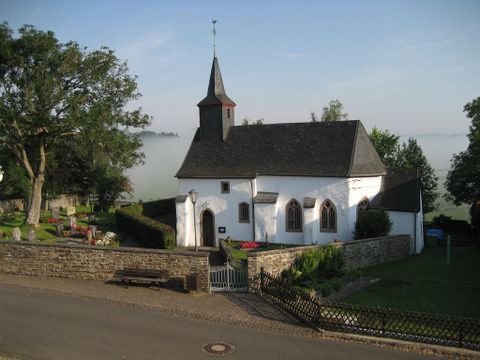 In der Mitte des bildes sieht man die kleine weißgetünchte Kapelle. Das Dach ist mit schwarzem Schiefer gedeckt. an der linken Seite ist ein ebenfalls schwarzgedeckter Kichturm mit spitzem Dach.
Auf der Kirchturmspitze sieht man einen Wetterhahn. Umgeben wird die Kapelle von einen Kirchhof, der von einer Bruchsteinmauer umrahmt ist.