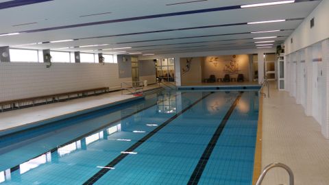 Innenansicht des Hallenbads mit einem langen Schwimmbecken, weißen Fliesenwänden und einem Sitzbereich im Hintergrund.