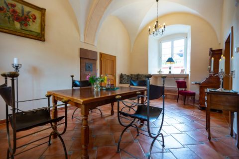 Ein helles Zimmer mit einem Holzstammtisch und metallenen Stühlen. Neben dem Fenster stehen Blumen und ein kleiner Tisch.