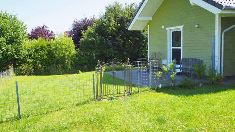 Ein kleines, grünes Haus mit einem gepflegten Garten. Das Grundstück ist von einem Zaun umgeben und es gibt viel Gras und Bäume in der Umgebung.