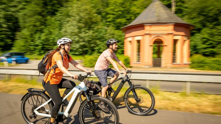 Ein Mann und eine Frau fahren auf E-Bikes entlang eines Radweges vorbei an dem großen Quelltempel der Lindenquelle bei Birresborn.