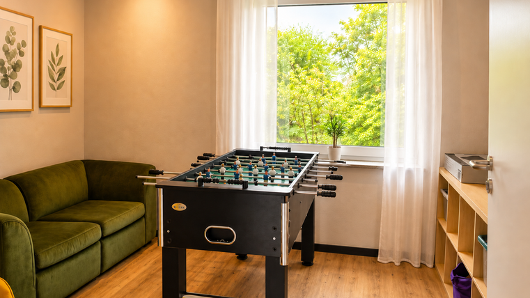 Une pièce confortable avec une table de baby-foot au milieu. De grandes fenêtres avec vue sur la verdure assurent beaucoup de lumière naturelle.