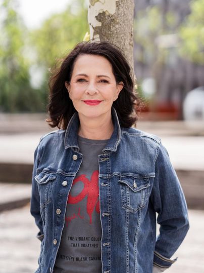 Eine Frau steht lächelnd vor einem Baum. Sie trägt eine Jeansjacke und ein T-Shirt mitdruck.