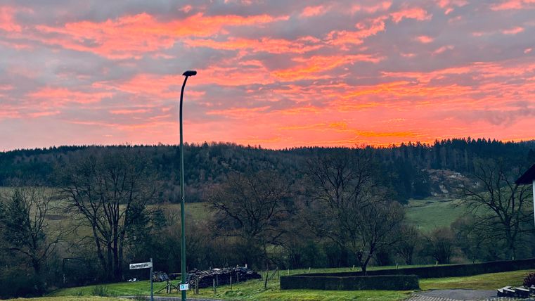 Ein malerischer Sonnenuntergang mit leuchtend roten und orangefarbenen Wolken. Im Vordergrund sind Bäume und eine ruhige Landschaft sichtbar.