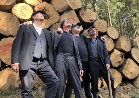 Gruppenbild der Musiker, wie sie arm im arm vor Holzstämmen stehen mit mit Blick in den Himmel