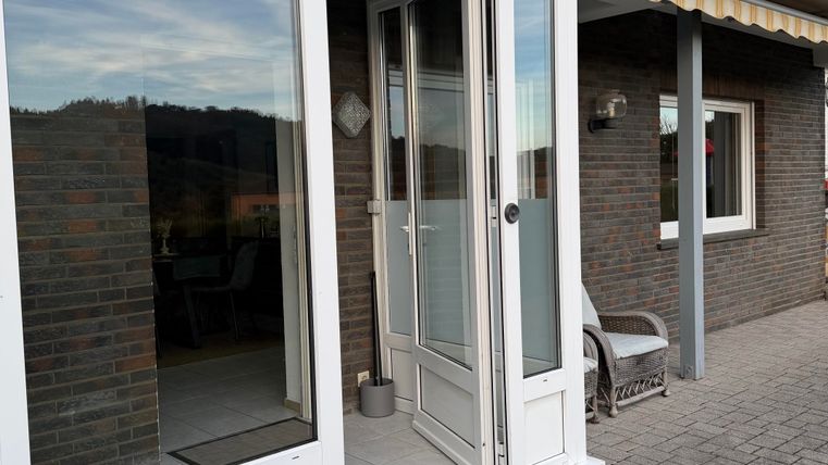 Un hall d'entrée avec une porte claire et de grandes fenêtres. À côté de la porte se trouve un fauteuil confortable et en arrière-plan, plusieurs fenêtres sont visibles.