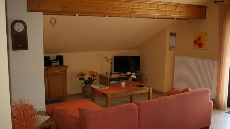 Ein gemütliches Wohnzimmer mit einem roten Sofa, einem Fernseher und einem Holztisch. Die Wände sind hell und die Dekoration ist einfach und freundlich.