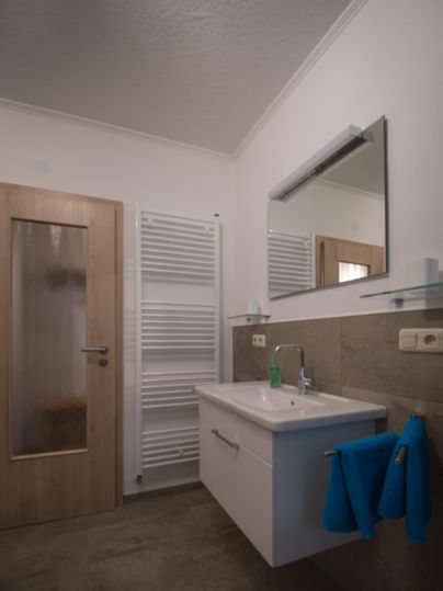 A modern bathroom with a sink, mirror, and towel rack. The walls are light and the floor has a neutral tone.