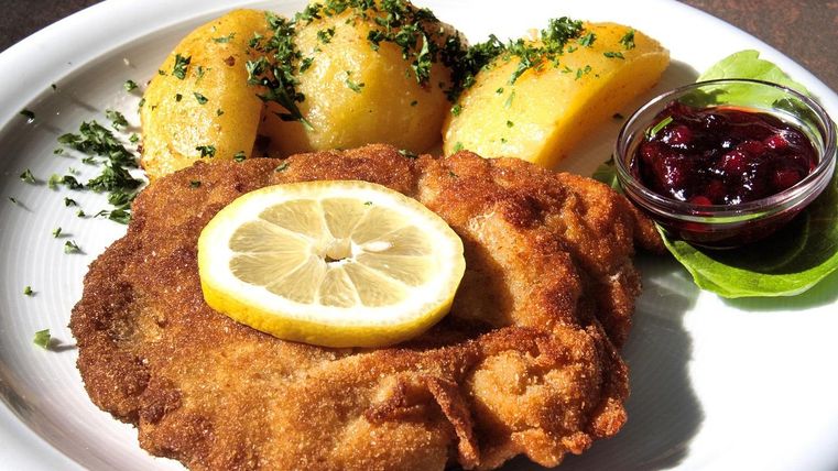 Ein Teller, auf dem ein großes paniertes Schnitzel mit einer Zitronenscheibe darauf liegt. Daneben drei Kartoffeln mit Kräutern und eine Schale mit Preiselbeeren darin.