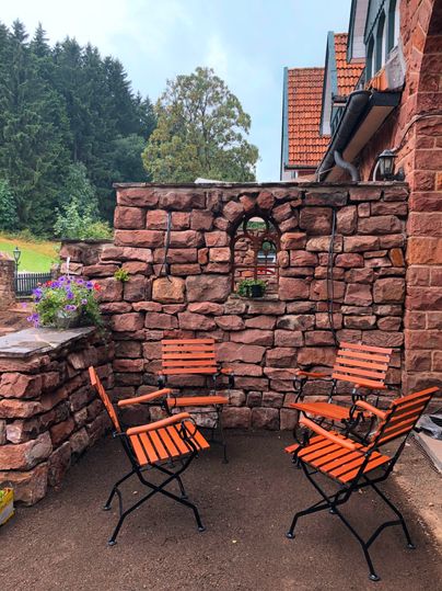 Un coin salon confortable avec des chaises orange devant un mur en pierre. En arrière-plan, on peut voir des arbres verts et un ciel dégagé.