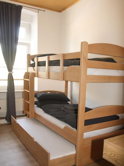 Ein einfaches Zimmer mit einem Holzstockbett und einem Ausziehbett. Die Wände sind hell, und ein Fenster lässt viel Tageslicht herein.