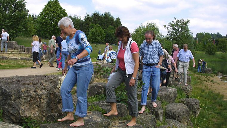 Menschen gehen barfuß über einen steinigen Pfad in einer grünen Parklandschaft. Der Himmel ist leicht bewölkt.