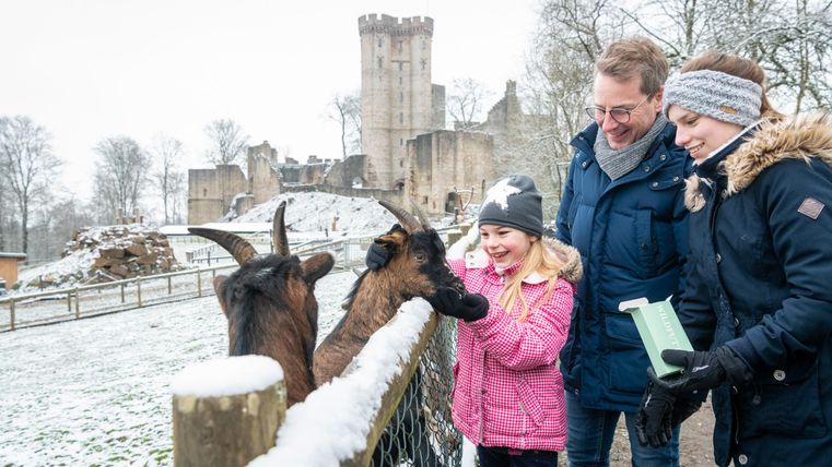 Eine Vater steht mit seinen zwei Töchtern an einem Zaun, über den sie Ziegen füttern. Die Wege und Felder sind verschneit. Im Hintergrund ragen die Türme einer Burg empor.