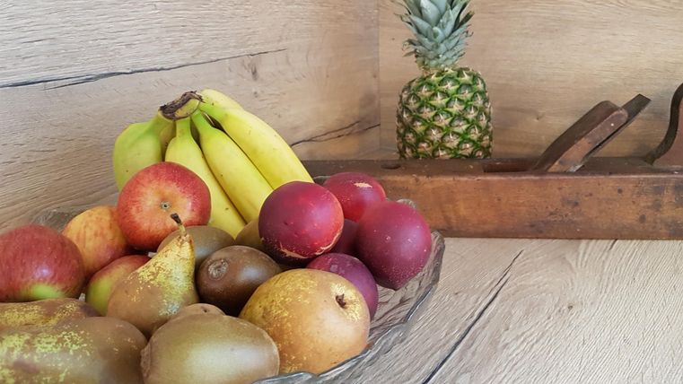 Ein Korb mit verschiedenen Früchten wie Äpfeln, Birnen, Bananen und Pflaumen. Im Hintergrund steht eine Ananas auf einem Holzregal.