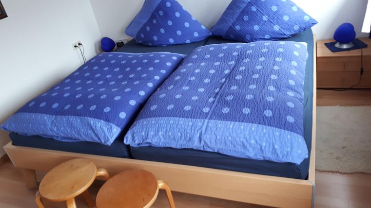 Ein gemütliches Schlafzimmer mit einem großen Doppelbett und blauer Bettwäsche. Neben dem Bett stehen zwei kleine Hocker aus Holz.