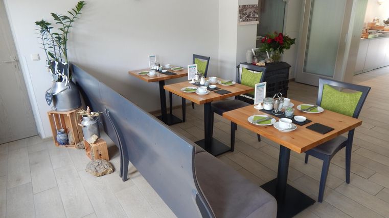 Ein modernes Restaurant mit holzfarbenen Tischen und stilvollen Stühlen. Die Atmosphäre ist freundlich und einladend.