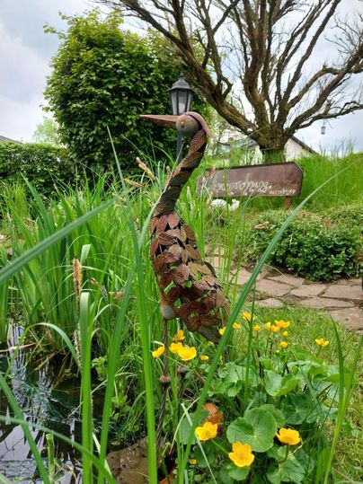 Een gestileerde reiger staat in een groene tuin met kleurrijke bloemen. Op de achtergrond zijn bomen en een pad zichtbaar.