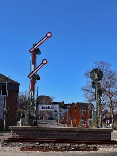 Een stationssignaal en verkeersveiligheidsborden staan in een rotonde. Op de achtergrond is een bord met de tekst "Gerolstein" te zien, en de lucht is helder en blauw.