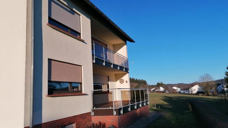 A modern residential building with several windows and balconies. In the background, a green meadow stretches out under a clear blue sky.