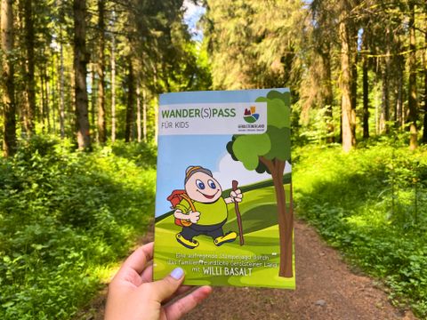 Das Prospekt "Wander(S)pass für Kids" wird hochgehalten mit einem dichten Wald und Waldweg im Hintergrund.