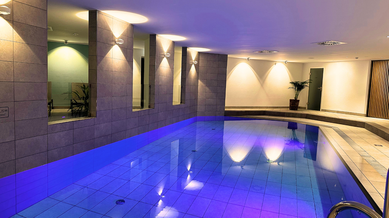 Ein modernes Schwimmbad mit leuchtendem Wasser und stilvollem Design. Die Wände sind mit fliesen verkleidet und es gibt gemütliche Beleuchtungselemente.