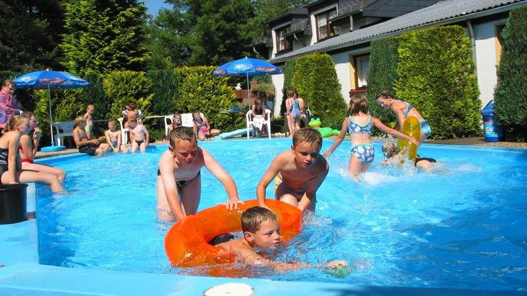 Ein beheizter Außenpool voller fröhlicher Kinder. Sie spielen und schwimmen, umgeben von grünen Hecken und Sonnenschirmen.