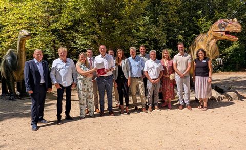 13 Personen stehen nebeneinander vor zwei großen Dinosaurier Stauten und lächeln glücklich in die Kamera. Zwei Personen in der Mitte halten eine Mappe hoch.