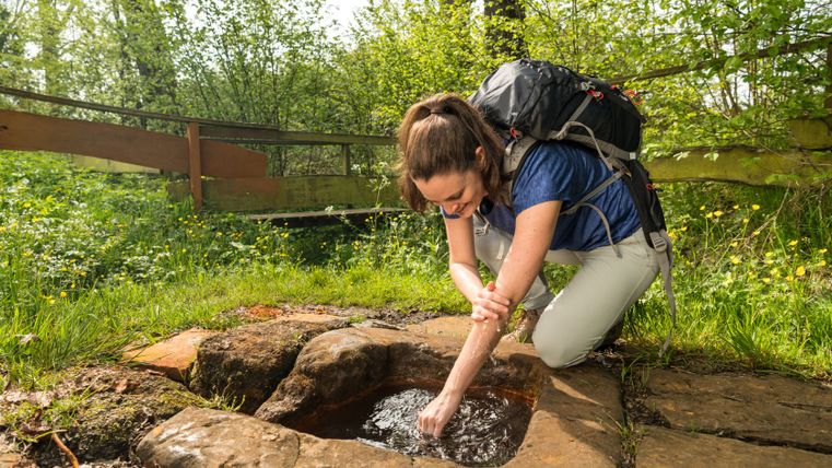 Eine Frau mit Rucksack erfrischt sich an einer natürlichen Quelle im Wald.