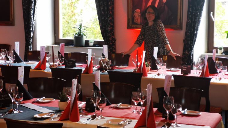 L'image montre une salle à manger élégamment décorée, probablement préparée pour un événement ou une célébration formelle. L'accent est mis sur plusieurs longues tables, recouvertes de nappes blanches et de chemins de table rouges. Chaque place comprend des couverts polis – fourchettes à droite des assiettes, couteaux derrière et cuillères complètement à gauche. Des serviettes rouges pliées en cônes triangulaires servent de centre de table sur chaque assiette.