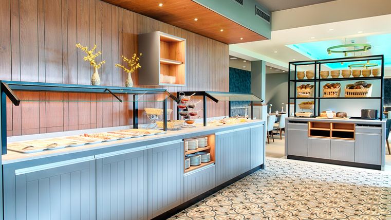 Ein modernes Buffet mit verschiedenen Speisen und einer eleganten, hellen Inneneinrichtung. Der Boden ist mit einem ansprechenden Muster gestaltet und es gibt dekorative Pflanzen.