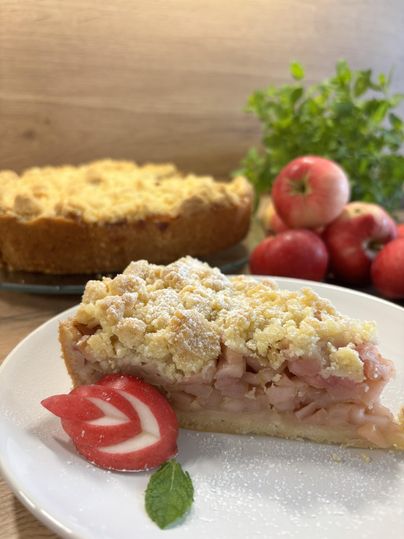Ein appetitliches Stück Apfel-Krümel-Kuchen liegt auf einem Teller. Im Hintergrund der Rest des Kuchens neben einem Haufen Äpfel.