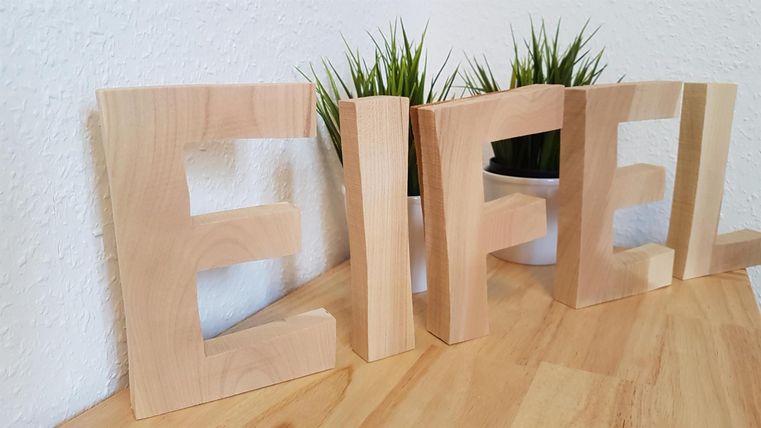 Holzbuchstaben bilden das Wort "Eifel". Im Hintergrund sind kleine Pflanzen sichtbar.