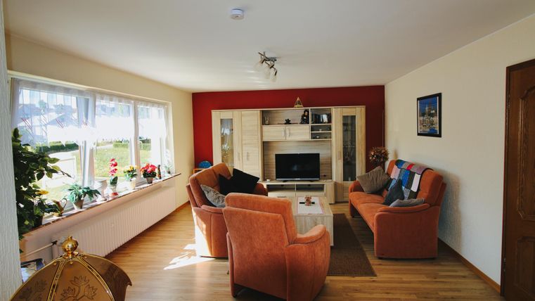 Ein gemütliches Wohnzimmer mit orangefarbenen Sofas und einem Fernseher. Große Fenster lassen viel Licht herein, und es gibt dekorative Pflanzen.