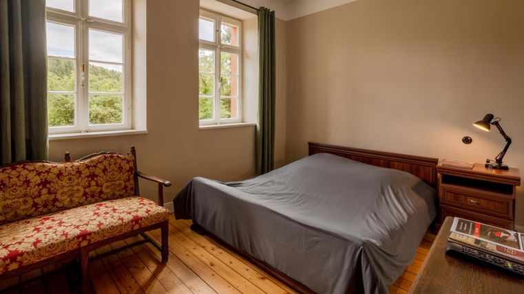 Ein einfaches Schlafzimmer mit einem Bett und einem kleinen Tisch. Es gibt große Fenster, die viel Tageslicht hereinlassen.