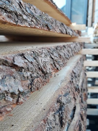 Ein Stapel von Brettern mit rauer Rinde, die Holzstruktur ist gut sichtbar. Im Hintergrund sind einige Holzpaletten zu erkennen.