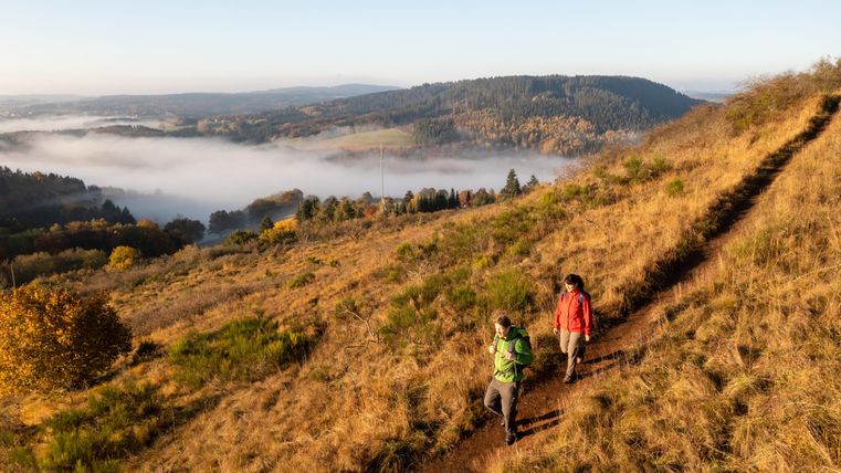 zwei Wanderer gehen auf einem schmalen Pfad einen Berg hinunter. Der Berg ist voller Laub und kahler Sträucher. Im Hintergrund weitere Berge und Nebel in den Tälern.