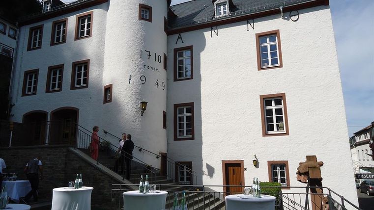 Weißes historisches Gebäude mit runden Türmen, datiert auf 1710 und 1949. Menschen stehen bei Stehtischen im Vordergrund.