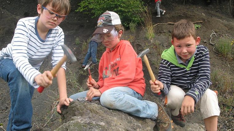 Drei Kinder mit Schutzbrillen und Hämmern sitzen auf einem Felsen. Sie tragen Freizeitkleidung und sind in einer natürlichen Umgebung.