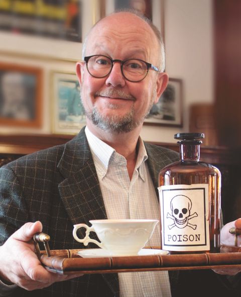 Ein lächelnder Mann mit Brille hält ein Tablett mit einer Teetasse und einer Flasche mit Totenkopf-Etikett, auf dem 'Poison' steht.