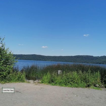 Ein schöner See mit klarem Wasser und grünen Pflanzen am Ufer. Im Hintergrund sind sanfte Hügel und ein blauer Himmel zu sehen.