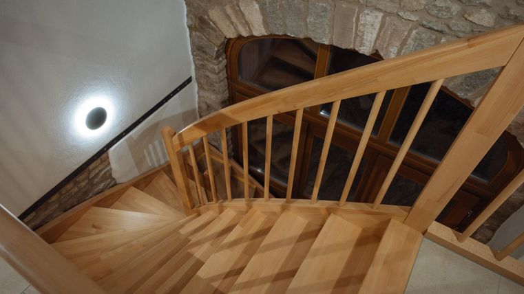Eine Holztreppe verläuft spiralförmig nach unten. Im Hintergrund sind steinerne Wände und ein gemütlicher Wohnbereich zu sehen.