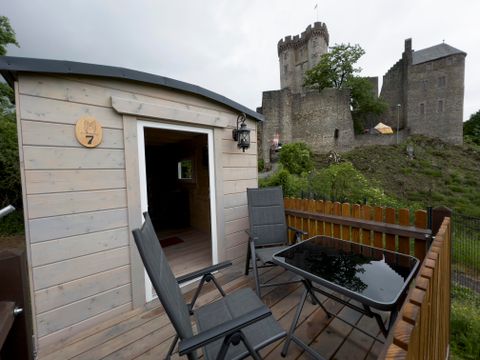 Ein gemütliches Häuschen mit einer Terrasse und zwei Stühlen. Im Hintergrund steht eine beeindruckende Burg.