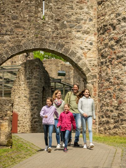 Zwei Eltern mit Ihren 3 Kindern laufen freudig durch einen massiven Torbogen der Kasselburg.