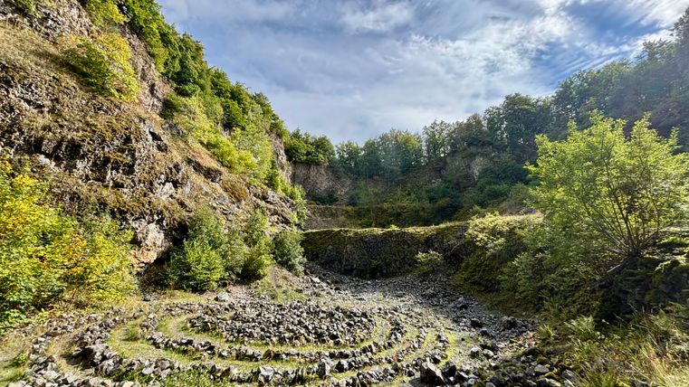 Steinspirale im Arensberg Vulkan mit bewaldeten Felswänden und blauem Himmel.