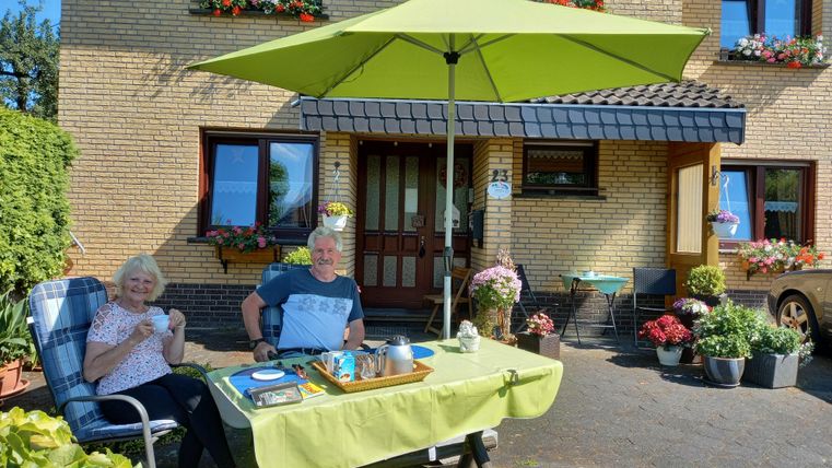 An older couple sits relaxed under a large green sun umbrella at a table. Around them, plants and flowers bloom in front of a cozy house.